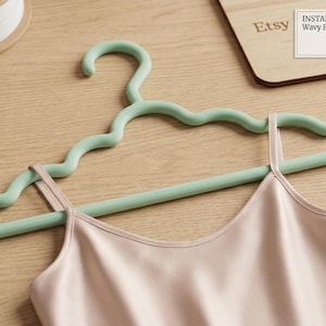 Puede incluir: Una percha ecológica ondulada verde claro con una camisola rosa pálido. La percha tiene un diseño ondulado único. La imagen también incluye una bobina de filamento blanco y un letrero de madera con el logotipo de Etsy y "Instant STL Download: Wavy Eco-Hanger File."
