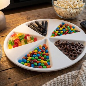 May include: A white divided serving platter filled with snacks. The platter contains colorful candies, gummy bears, chocolate pretzels, and black licorice. Two glass bowls of popcorn are also visible, along with a remote control.
