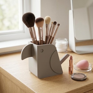 Può includere: Un porta pennelli per trucco grigio a forma di uccello con diversi pennelli per trucco all'interno. Un rossetto, una spugnetta per il trucco e un ombretto compatto sono sulla superficie di legno. Il supporto ha un becco e un occhio circolare.