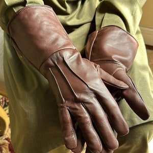 May include: A pair of brown leather gloves with a long cuff. The gloves have a smooth leather finish and are folded in half.