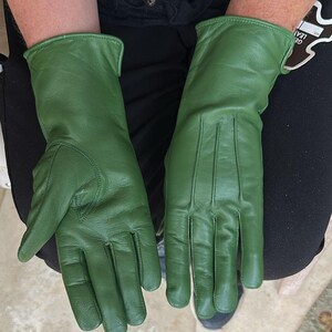 May include: A pair of green leather gloves. The gloves are a solid, vibrant green color. They have a classic design with a smooth texture and visible stitching details. The gloves are shown on a person's hands.