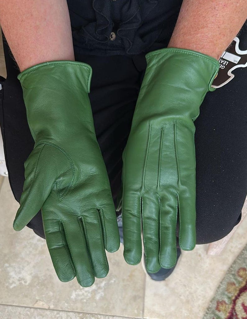 May include: A pair of green leather gloves. The gloves are a solid, vibrant green color. They have a classic design with a smooth texture and visible stitching details. The gloves are shown on a person's hands.