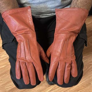 May include: A pair of reddish-brown leather gloves. The gloves have a gauntlet style cuff and are designed with multiple seams for flexibility. The leather appears to be slightly textured.