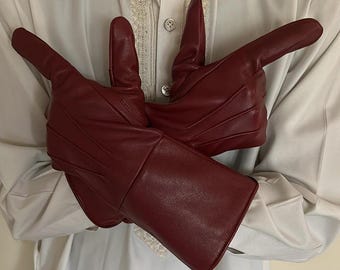 May include: A pair of burgundy leather gloves with a gauntlet style cuff. The gloves are a deep red color and appear to be made of a smooth leather material. The gloves are held up in front of a person wearing a white shirt.
