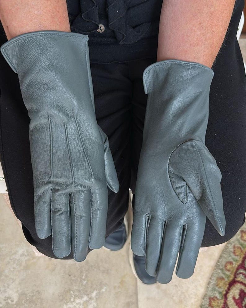 May include: A pair of gray leather gloves. The gloves have a classic design with a smooth texture and visible stitching details. The gloves are long, extending past the wrist.