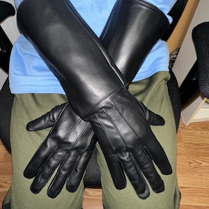 May include: A pair of black leather gloves with long gauntlets. The gloves are made of smooth, shiny leather and are designed to cover the hands and forearms. The gloves are resting on a person's lap, with the fingers of the gloves spread out.