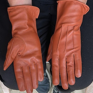 May include: A pair of rust-colored leather gloves. The gloves have a classic design with stitched details on the fingers and a cuff at the wrist. The gloves are shown on a person's lap, with black pants and blue sneakers visible.