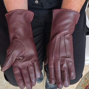 May include: A pair of burgundy leather gloves. The gloves are a deep red-purple color, with a smooth texture and visible stitching details. The gloves have a classic design, with a long cuff.