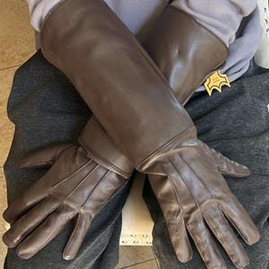 May include: Brown leather gloves with long sleeves. The gloves are a solid brown color and appear to be made of a smooth leather material. The gloves have a tag attached to the sleeve.
