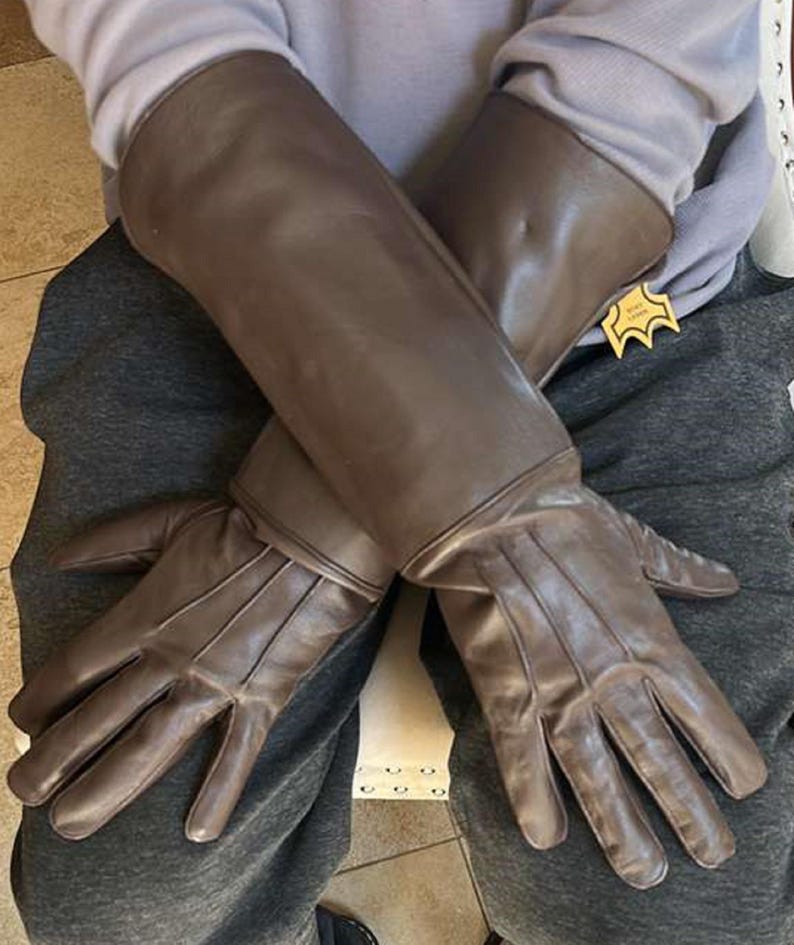 May include: Brown leather gloves with long sleeves. The gloves are a solid brown color and appear to be made of a smooth leather material. The gloves have a tag attached to the sleeve.