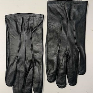 May include: A pair of black leather gloves featuring three vertical stitched lines on the back of each hand. A small white tag attached to the top of one glove displays the text Genuine Leather Quality in a decorative shape.
