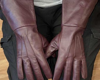 May include: A pair of burgundy leather gloves with long cuffs. The gloves have a classic design with visible stitching and a smooth texture. The gloves are designed to cover the hands and wrists.