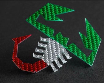 Può includere: Un emblema a forma di scorpione verde, bianco e rosso con un motivo in fibra di carbonio. L'emblema è realizzato in un materiale lucido e riflettente.