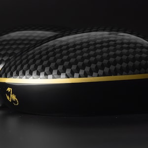 Op de afbeelding: Twee zwarte en gouden carbon fiber golfclub-hoofdbedekkingen met een schorpioenlogo.