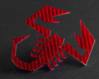 Può includere: Un emblema rosso a forma di scorpione con un motivo in fibra di carbonio. L'emblema è realizzato in un materiale sottile e flessibile ed è progettato per essere fissato a una superficie.