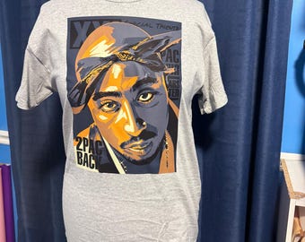 May include: A gray t-shirt with a black and white illustration of rapper 2Pac on the front. The illustration is in a pop art style and features 2Pac wearing a bandana. The text "2PAC BACK" is printed below the illustration. The text "SPECIAL TRIBUTE" is printed above the illustration.