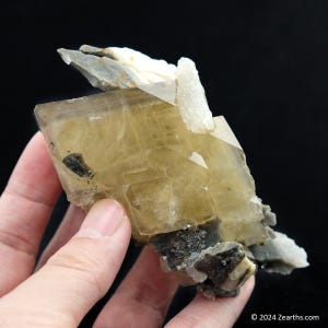 Puede incluir: Un gran cristal de calcita transparente, de color amarillo-marrón, con múltiples facetas. El cristal está unido a una matriz más pequeña, gris y blanca.