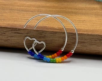 May include: Rainbow beaded hoop earrings with two silver heart charms. The earrings are made with small, colorful beads in a rainbow pattern.