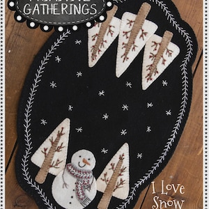 May include: Black oval table mat with a snowman and four trees. The mat has a white stitched border and snowflake accents. The text "I Love Snow Table Mat" is displayed. Finished size: 12" x 18".