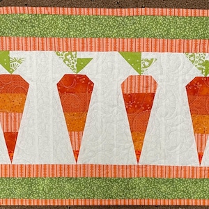 Op de afbeelding: Een rechthoekige, gewatteerde tafelloper met zes gestileerde oranje wortels met groene toppen op een witte achtergrond. De loper is omrand met groen en oranje patroonstof, wat een levendige, handgemaakte uitstraling geeft. Geschikt voor lente- of paasdecoratie.