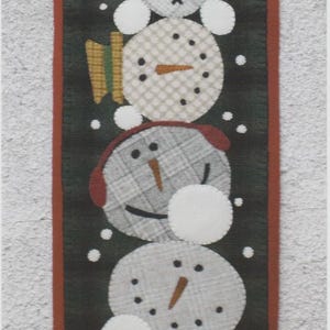 May include: A vertical wool applique featuring a series of snowman faces in various patterns and colors, with a dark green background and a red border. The dimensions are 11" x 40.5".