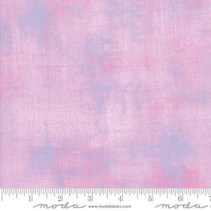 May include: A fabric with a textured, painterly design. The background is a soft pink, with patches of lavender and deeper pink hues. The fabric has a subtle, brushed appearance, and a ruler is at the bottom.