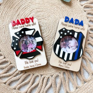 May include: Two wooden photo frames with the words "DADDY" and "DADA" in red and blue, respectively. Each frame has a circular photo opening and the text "Please come home safe!" and "We love you!" with names. One frame is shaped like a firefighter's emblem.