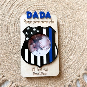 May include: Wooden photo frame with a police badge design. The frame features the word "DADA" in blue letters at the top, with the message "Please come home safe!" below. The frame includes a photo of two children and the words "We love you! Mama & Masen."