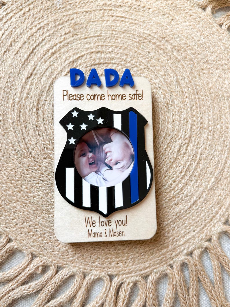 May include: Wooden photo frame with a police badge design. The frame features the word "DADA" in blue letters at the top, with the message "Please come home safe!" below. The frame includes a photo of two children and the words "We love you! Mama & Masen."