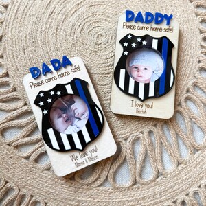 May include: Two wooden photo frames with a police badge design. One says "DADA Please come home safe!" and the other says "DADDY Please come home safe!". Each frame has a photo of a baby. One says "I love you! Brixton" and the other says "We love you! Mama & Masen".