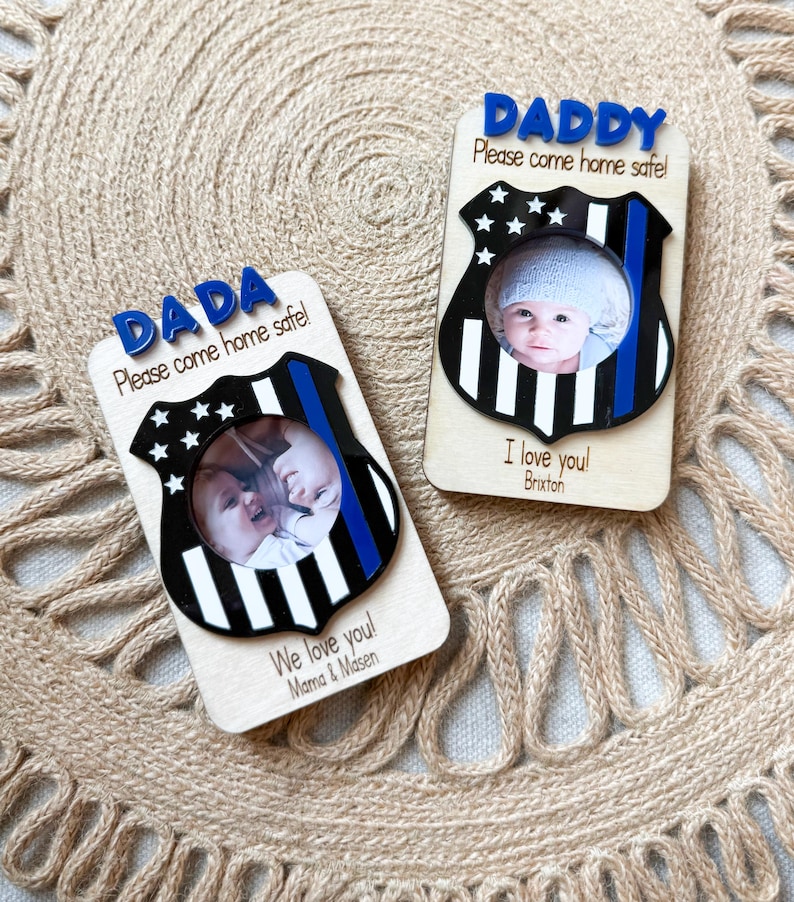 May include: Two wooden photo frames with a police badge design. One says "DADA Please come home safe!" and the other says "DADDY Please come home safe!". Each frame has a photo of a baby. One says "I love you! Brixton" and the other says "We love you! Mama & Masen".
