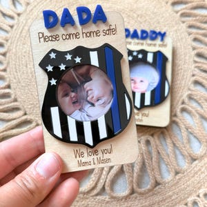 May include: Wooden photo frame with a police badge design. The frame has the word "DADA" in blue letters at the top, with the message "Please come home safe!" and "We love you! Mama & Masen" printed on it. The frame holds a circular photo.