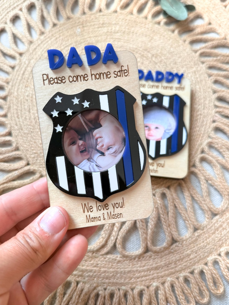 May include: Wooden photo frame with a police badge design. The frame has the word "DADA" in blue letters at the top, with the message "Please come home safe!" and "We love you! Mama & Masen" printed on it. The frame holds a circular photo.