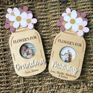 Pode incluir: Duas molduras de fotos de madeira em forma de pote Mason. Cada moldura tem um arranjo floral no topo em branco, rosa e dourado. Uma moldura diz "Vovó" e a outra diz "Mamãe". As molduras incluem o texto "Flower's For" e os nomes das pessoas que as amam.