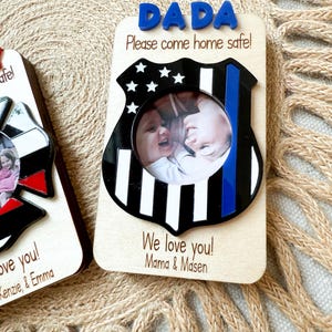 May include: Wooden photo frame with a police badge design. The frame has the word "DADA" in blue letters at the top, with the message "Please come home safe!" below. The frame includes a photo of two children and the message "We love you! Mama & Masen."