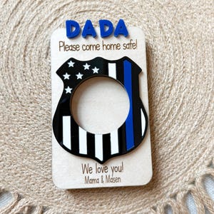 May include: A wooden plaque with the word "DADA" in blue letters at the top. Below, it reads "Please come home safe!" and features a black and white police badge design with a blue stripe. The bottom reads "We love you! Mama & Masen."