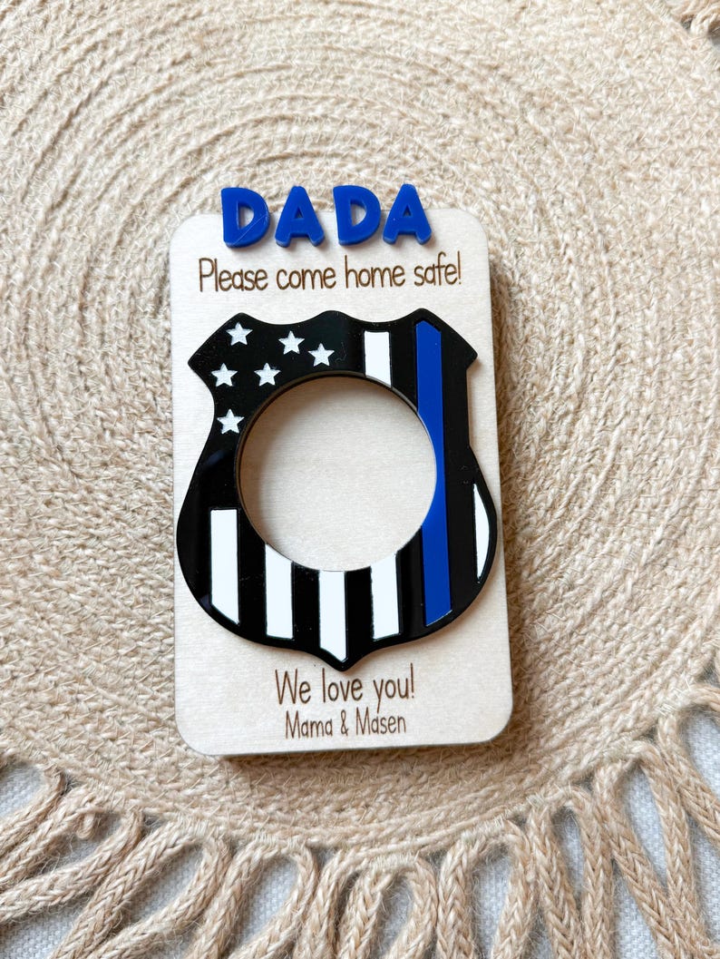 May include: A wooden plaque with the word "DADA" in blue letters at the top. Below, it reads "Please come home safe!" and features a black and white police badge design with a blue stripe. The bottom reads "We love you! Mama & Masen."