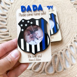 May include: Wooden photo frame with a police badge design. The frame has the word "DADA" in blue letters at the top, with the message "Please come home safe!" below. The frame includes a photo and the words "We love you! Mama & Masen."