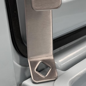 May include: A brushed stainless steel metal hook with a flat, vertical body and a curved top section. The base of the hook features a square-shaped cutout, and the entire piece has a smooth, metallic finish with a consistent, uniform texture.
