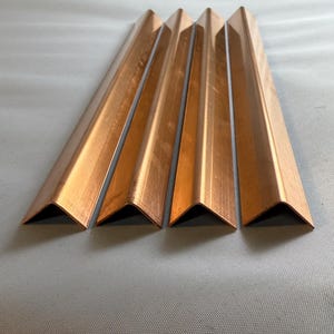 May include: Four copper corner trim pieces, each with a 90-degree angle, are arranged in a row on a light gray surface.