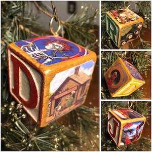 May include: Wooden Christmas ornament shaped like a block, with the letter "D" in red. The block features various images, including a skeleton with a floral design, a building, and other colorful graphics. The ornament hangs from a gold-colored hook.