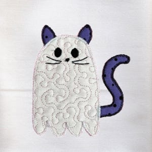 Peut inclure: Serviette de cuisine blanche avec un motif de chat fantôme brodé. Le chat a des oreilles, une queue et un contour violets, avec des yeux et des moustaches noirs. Le chat fantôme est matelassé avec du fil blanc. La serviette est pliée sur une planche à découper en bois.