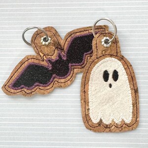 May include: Two Halloween-themed keychains. One is a bat with black wings and purple accents, the other is a white ghost. Both are made of cork material and have silver key rings.