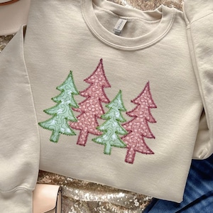 Könnte beinhalten: Cremefarbenes Sweatshirt mit einem gestickten Design von vier Weihnachtsbäumen. Die Bäume sind in Rosa- und Grüntönen mit grünen und roten Nähten gehalten. Das Sweatshirt hat einen Rundhalsausschnitt und lange Ärmel.