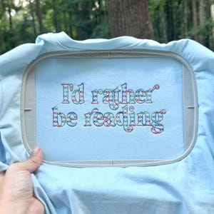 Peut inclure: Un t-shirt bleu clair fixé dans un tambour à broder en plastique gris. Le tissu présente le texte brodé "I'd rather be reading" dans une police à motifs multicolores avec des nuances de rouge, de vert et de bleu sur toutes les lettres.