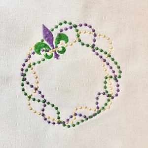 May include: A white fabric with an embroidered design of a purple fleur-de-lis surrounded by a wreath of green and gold beads.