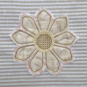 May include: A beige fabric flower with a pink outline and a yellow centre. The flower is embroidered on a striped fabric background.