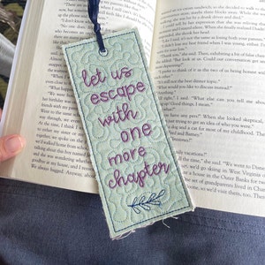 May include: A blue and green quilted bookmark with the text "Let us escape with one more chapter" in purple embroidery. The bookmark has a leafy design at the bottom.