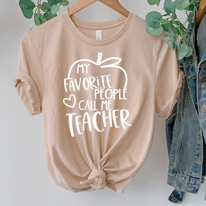May include: A tan t-shirt with the text "My Favorite People Call Me Teacher" in white, featuring an apple design. A denim jacket and a straw hat hang on a clothing rack, with greenery draped above.