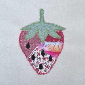 Puede incluir: Una aplicación de fresa de tela con un diseño de patchwork. La fresa presenta un tallo y una hoja verdes, con un cuerpo rojo y rosa. Se ven detalles de semillas negras. La aplicación está cosida sobre un fondo de tela beige claro.